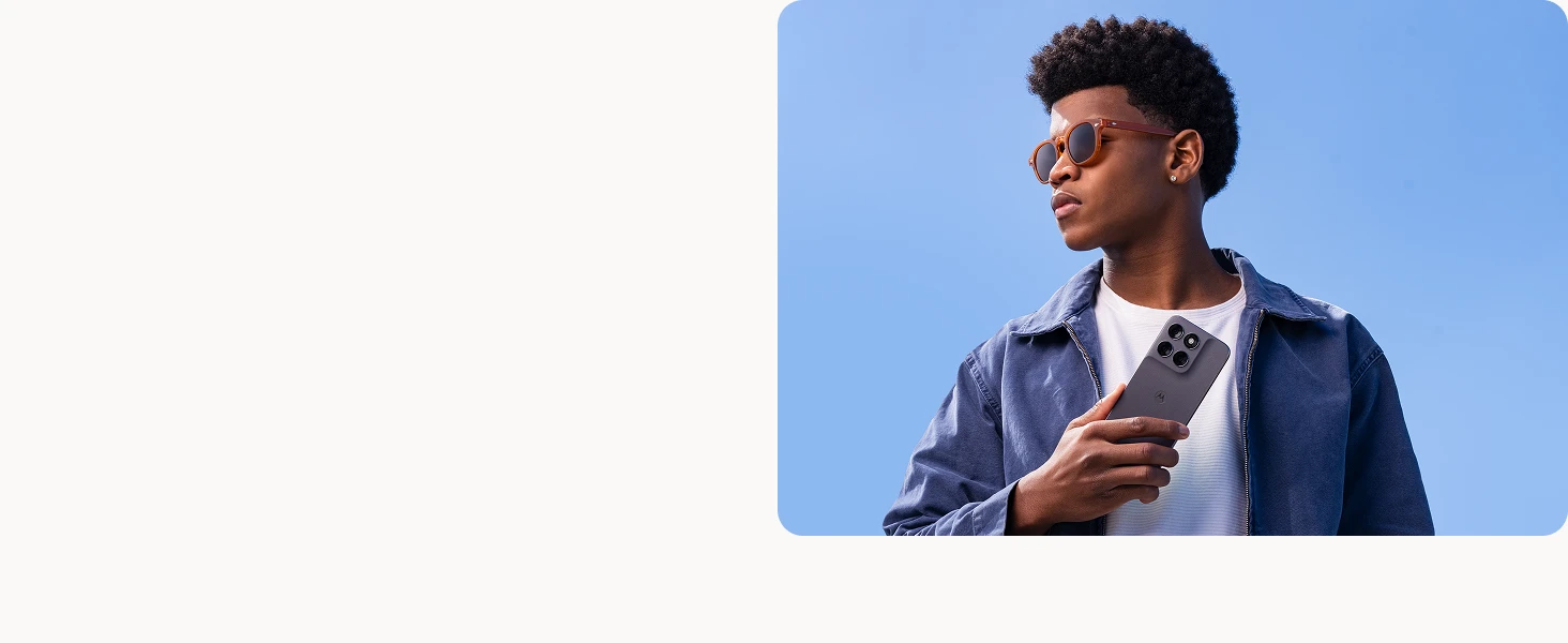 A young man wearing sunglasses and a blue jacket holds a gray smartphone against a clear blue sky background.