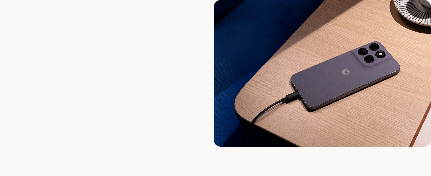 A smartphone with triple rear cameras is charging on a wooden table beside a blue chair and a cup on a saucer.