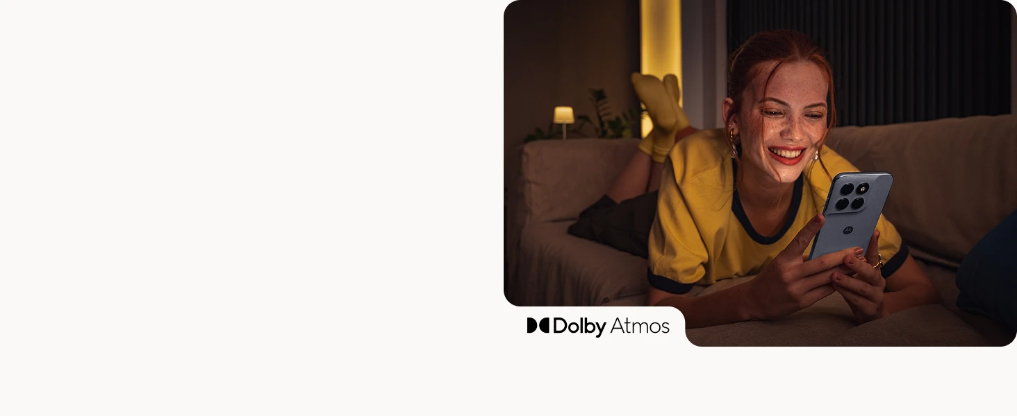 A smiling young woman with red hair lies on a couch looking at her smartphone, with Dolby Atmos branding in a warmly lit room.