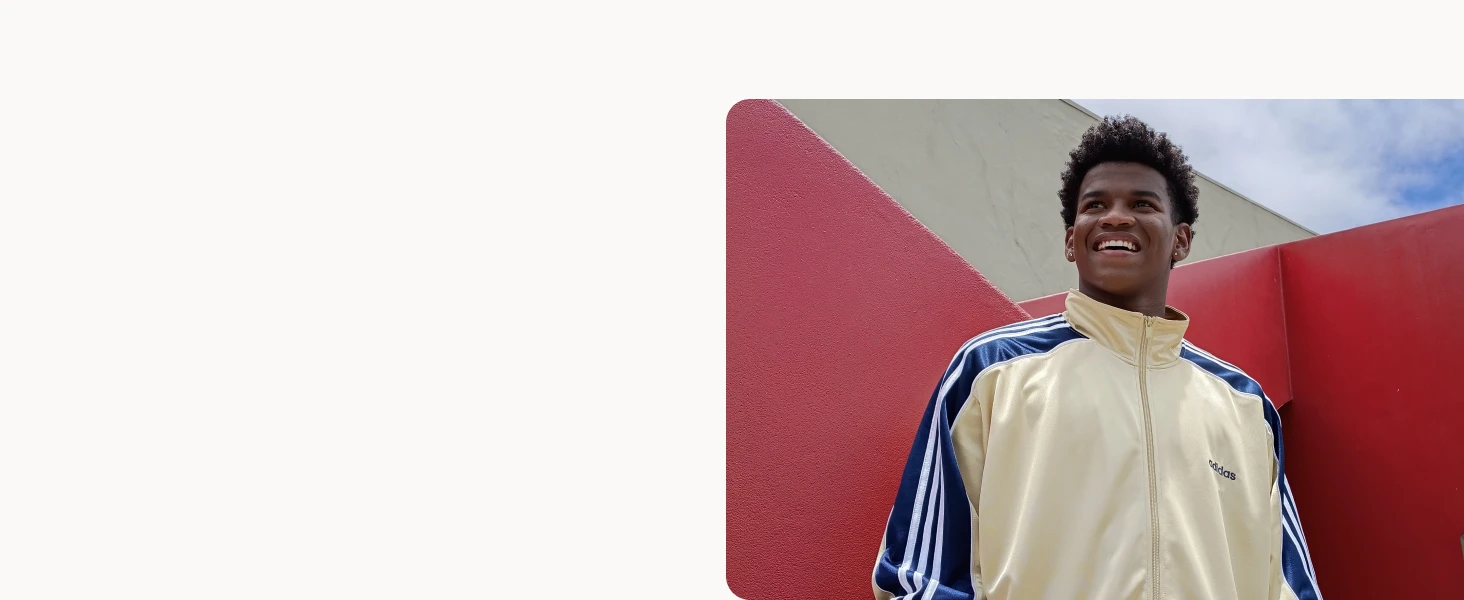 A young man smiles happily wearing a cream and navy Adidas jacket, standing outdoors by a red geometric structure with a partly cloudy sky in the background.