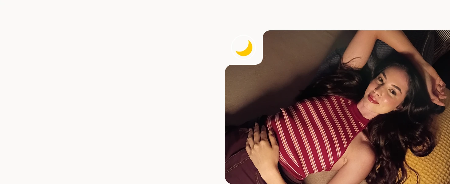 A young woman lies on a cozy surface with long dark hair, wearing a maroon striped sleeveless top, looking relaxed and comfortable.